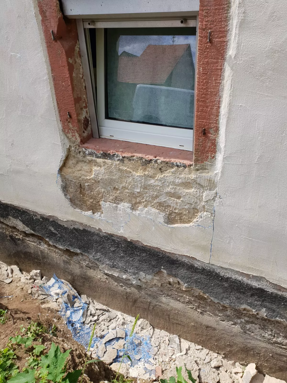 Ein kleines Fenster mit weißem Rahmen befindet sich in einer Wand mit beschädigtem Putz, der den Blick auf darunterliegende Ziegel freigibt. Die Wand weist Anzeichen von Verfall auf, mit freiliegendem Geflecht und abblätternden Schichten. Darunter liegen Trümmer auf dem Boden verstreut, darunter Putzstücke und ein Stück freiliegendes Fundament.