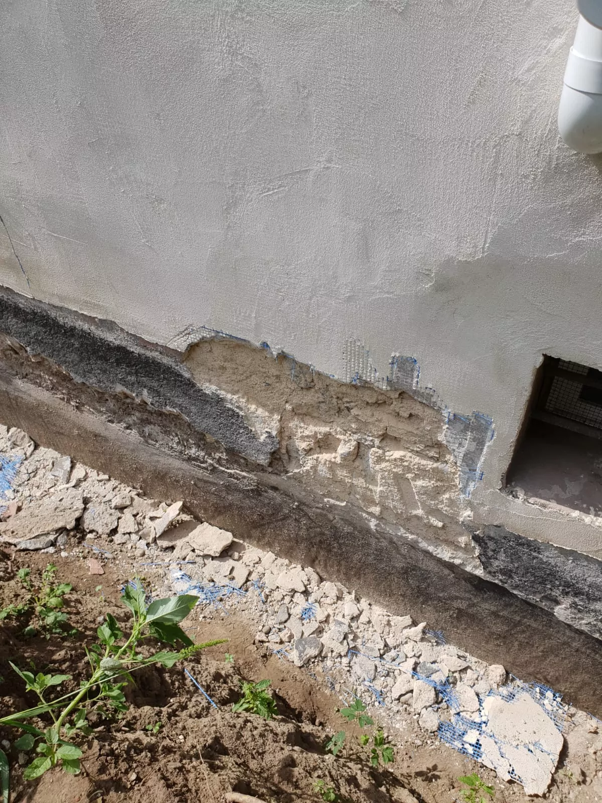 Das Bild zeigt einen beschädigten Abschnitt der Außenwand eines Gebäudes mit Rissen und abblätterndem Putz, der das darunterliegende Material freilegt. Trümmerteile liegen auf dem Boden und auf der rechten Seite befindet sich ein Abflussrohr. In der Nähe wächst eine kleine Pflanze.