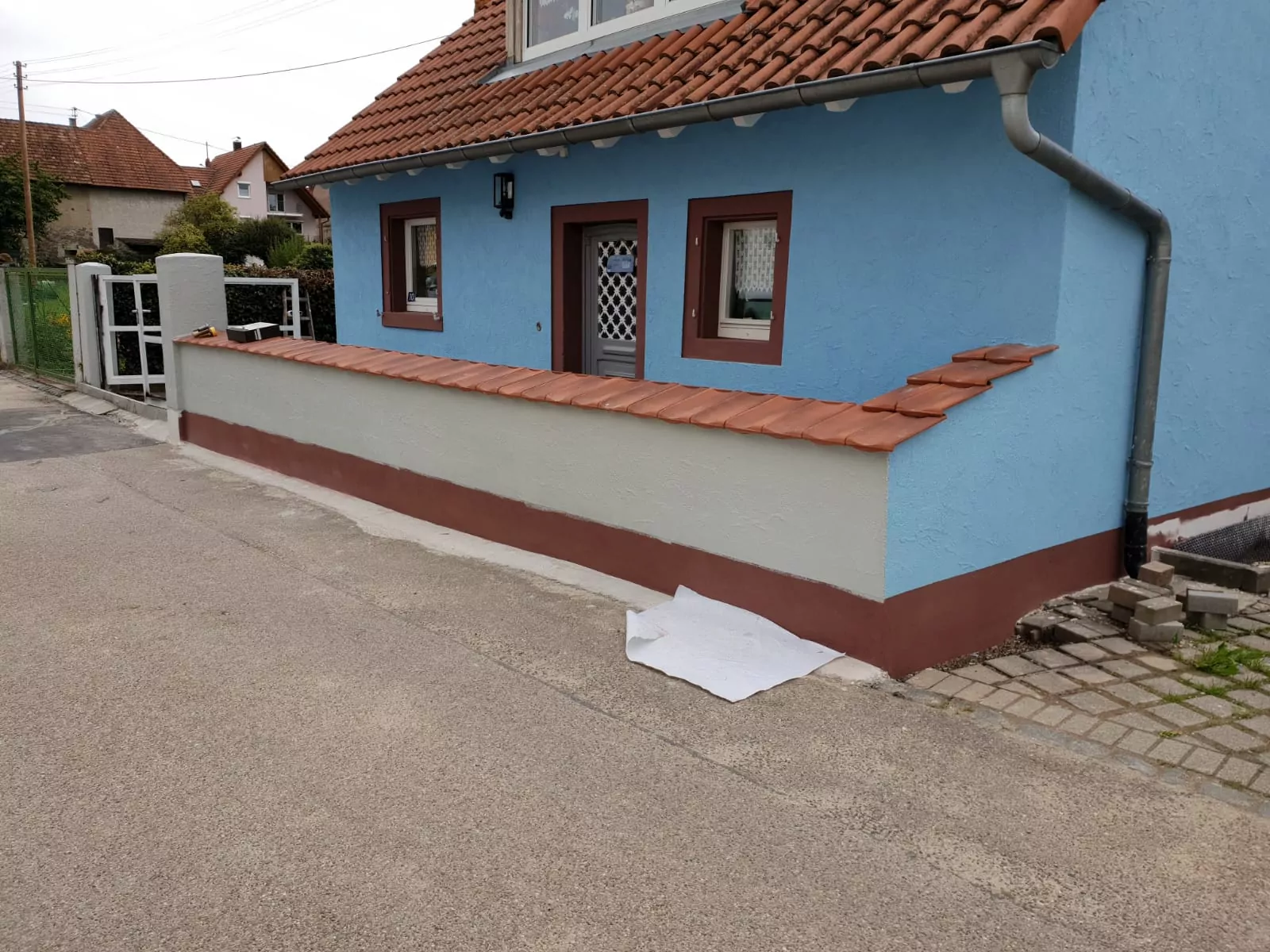 Parallel zur Straße verläuft ein blaues Haus mit rotem Ziegeldach und einer niedrigen angrenzenden Mauer. Die Mauer ist mit ähnlichen roten Ziegeln bedeckt. Auf dem Bürgersteig liegt ein zerknülltes weißes Laken, und links ist ein Metalltor zu sehen.