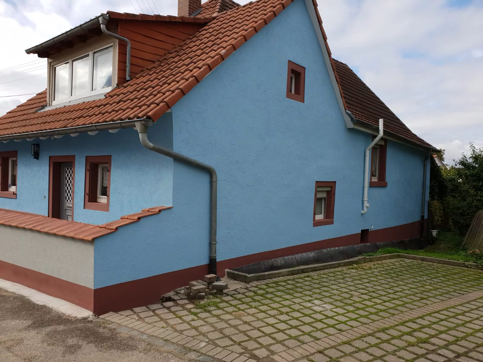 Ein kleines Haus mit blauen Wänden und einem braunen Ziegeldach. Es hat mehrere Fenster und eine graue Dachrinne. Im Vordergrund sind eine gepflasterte Auffahrt und ein Teil einer gepflasterten Straße zu sehen. Der Himmel ist bewölkt.