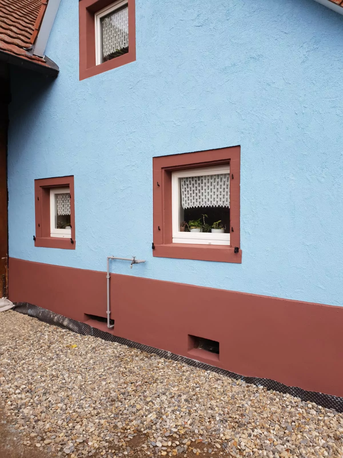 Ein Haus von außen mit blauer Oberwand und braunem Unterteil. Zwei kleine Fenster sind mit weißen Spitzenvorhängen versehen. Der Boden ist mit Kies bedeckt und unten ist eine Entlüftung sichtbar. Zwischen den Fenstern verläuft vertikal ein Metallrohr.