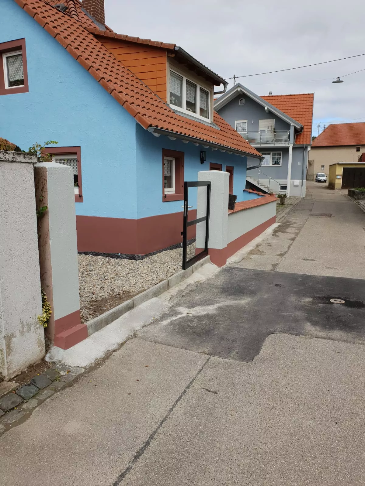 Straßenansicht eines kleinen Hauses mit blauen und roten Wänden und einem roten Dach. Das Haus hat einen Eingang mit einem Metalltor. Ein schmaler Gehweg verläuft neben dem Haus und im Hintergrund sind angrenzende Gebäude sichtbar.