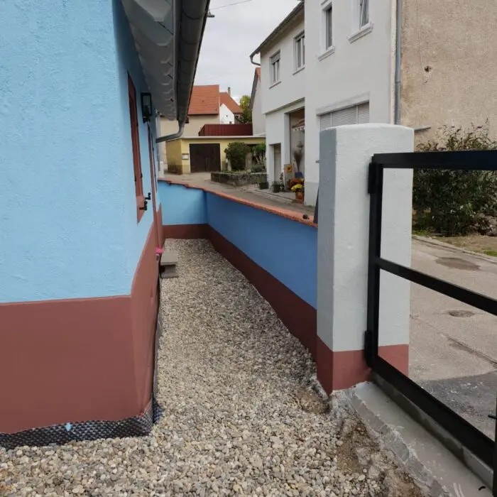 Ein schmaler Gehweg mit Kies verläuft neben einem blau-braunen Haus, das an eine weiß-braune Wand grenzt. Im Hintergrund sind weitere Häuser in einer ruhigen Straße zu sehen. Auf der rechten Seite ist teilweise ein schwarzes Tor zu sehen.