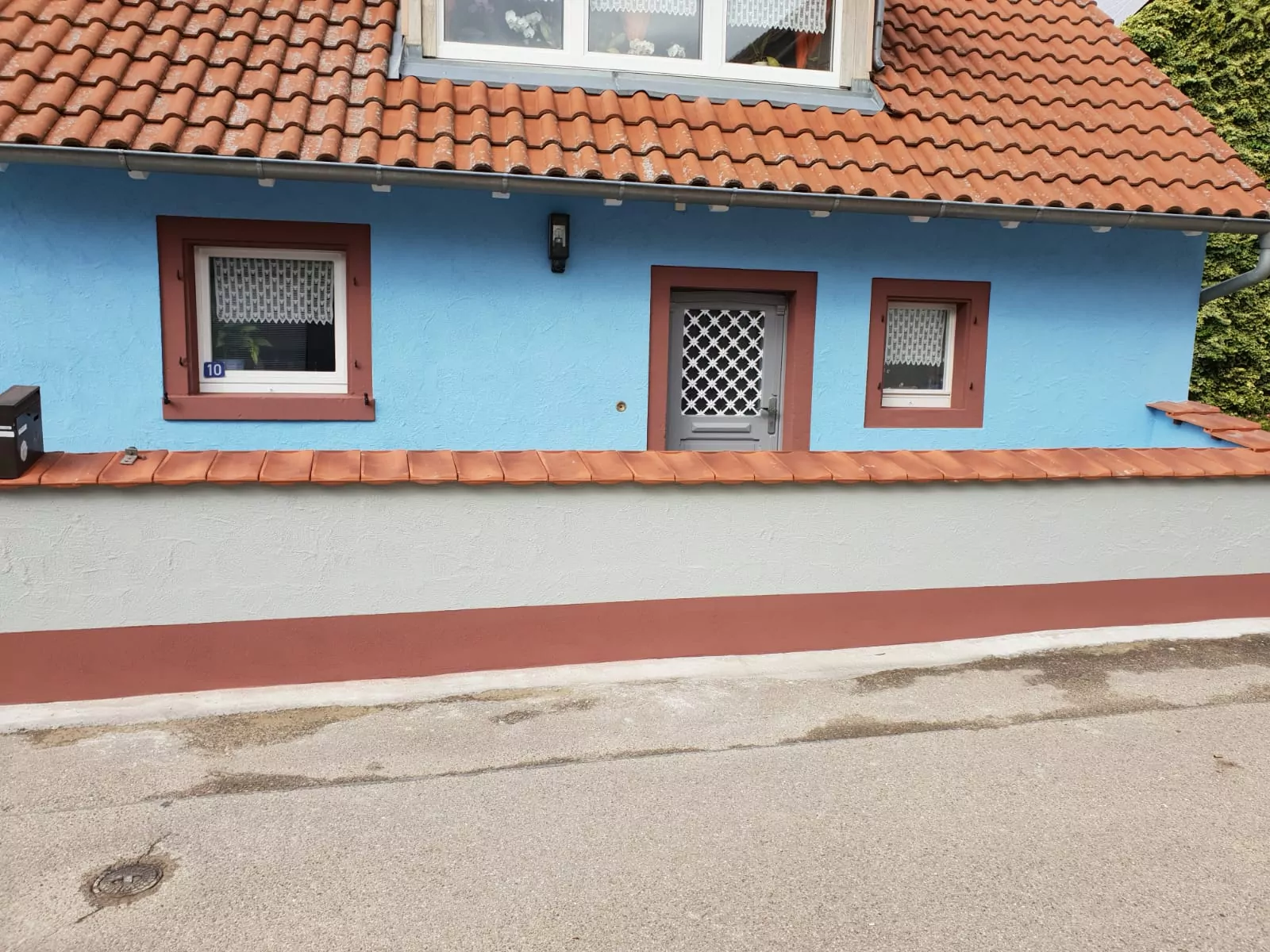 Ein kleines blaues Haus mit einem roten Ziegeldach und zwei Fenstern auf beiden Seiten einer grauen Tür, alle mit dekorativen weißen Spitzenvorhängen. Das Haus ist von einer niedrigen Mauer mit einer passenden roten Ziegelabdeckung umgeben. Die Straße davor ist gepflastert.