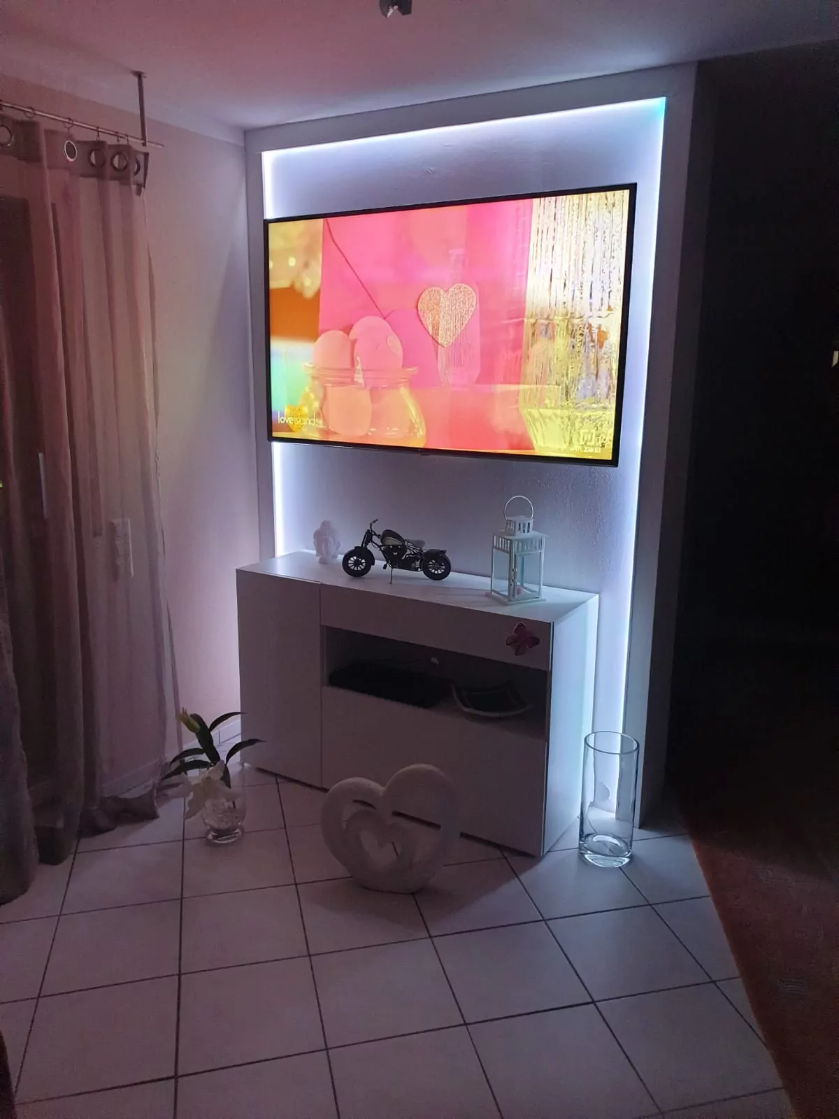 Ein an der Wand montierter Fernseher zeigt ein farbenfrohes, abstraktes Herzbild. Darunter befindet sich ein weißer Schrank mit einem kleinen Motorradmodell und einer Laterne. Auf dem Boden liegt ein herzförmiges Ornament. Der Raum verfügt über geflieste Böden, eine beleuchtete Kulisse und transparente Vorhänge am Fenster.