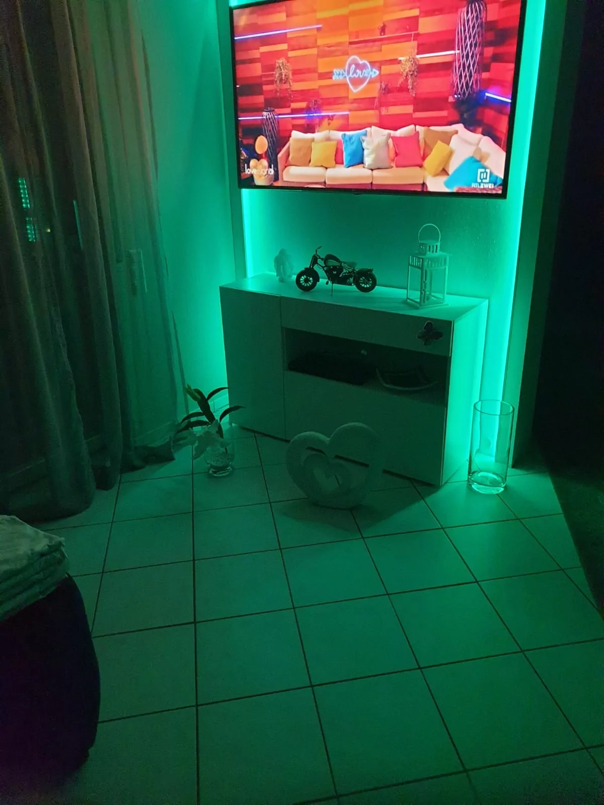 In einem Raum mit leuchtendem grünem Licht steht ein Fernseher, auf dem ein buntes Programm läuft, möglicherweise eine Talkshow. Darunter steht ein weißer Schrank mit einem dekorativen Motorradmodell, außerdem stehen eine Bodenvase und eine Pflanze. Der Boden des Raums ist weiß gefliest.