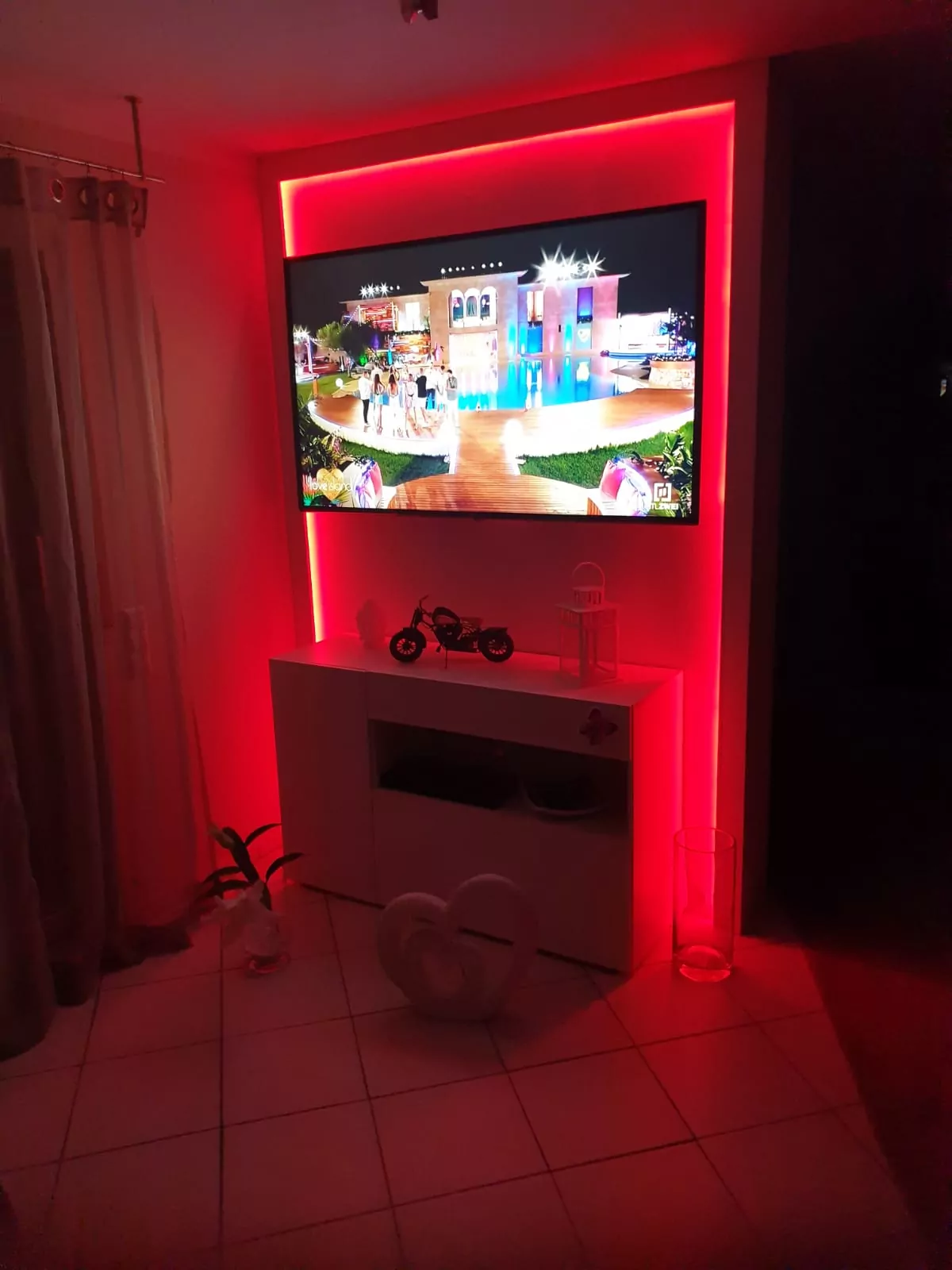 Ein an der Wand montierter Fernseher zeigt eine lebendige Szene, umgeben von rosa Hintergrundbeleuchtung. Darunter hält eine weiße Konsole ein Miniaturmotorrad und Dekorationsgegenstände. Auf dem Boden befindet sich ein herzförmiges Ornament und links sind Vorhänge.