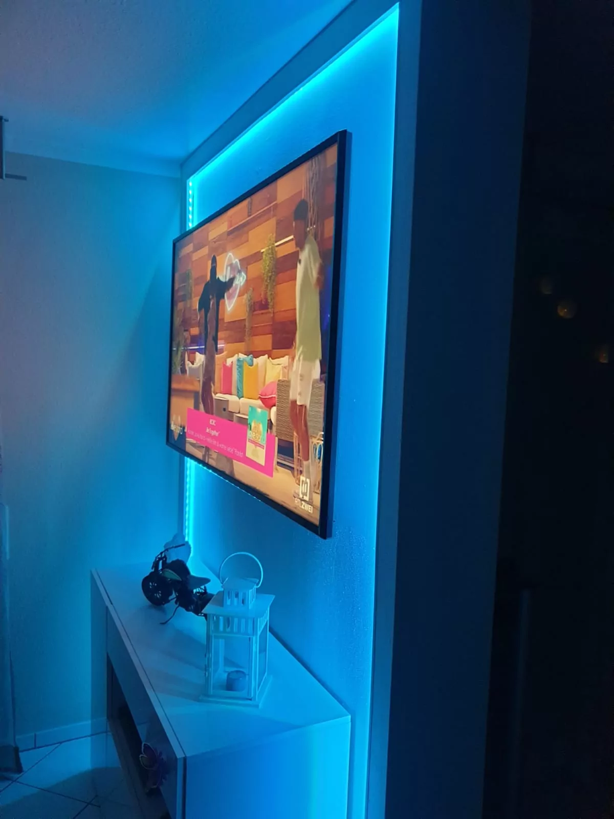 Ein an der Wand montierter Fernseher zeigt eine Szene mit zwei Personen in einem hell erleuchteten Raum. Der Fernseher ist von einer blauen LED-Hintergrundbeleuchtung umgeben. Unter dem Fernseher befindet sich ein Regal mit einer dekorativen Laterne und einigen elektronischen Geräten.