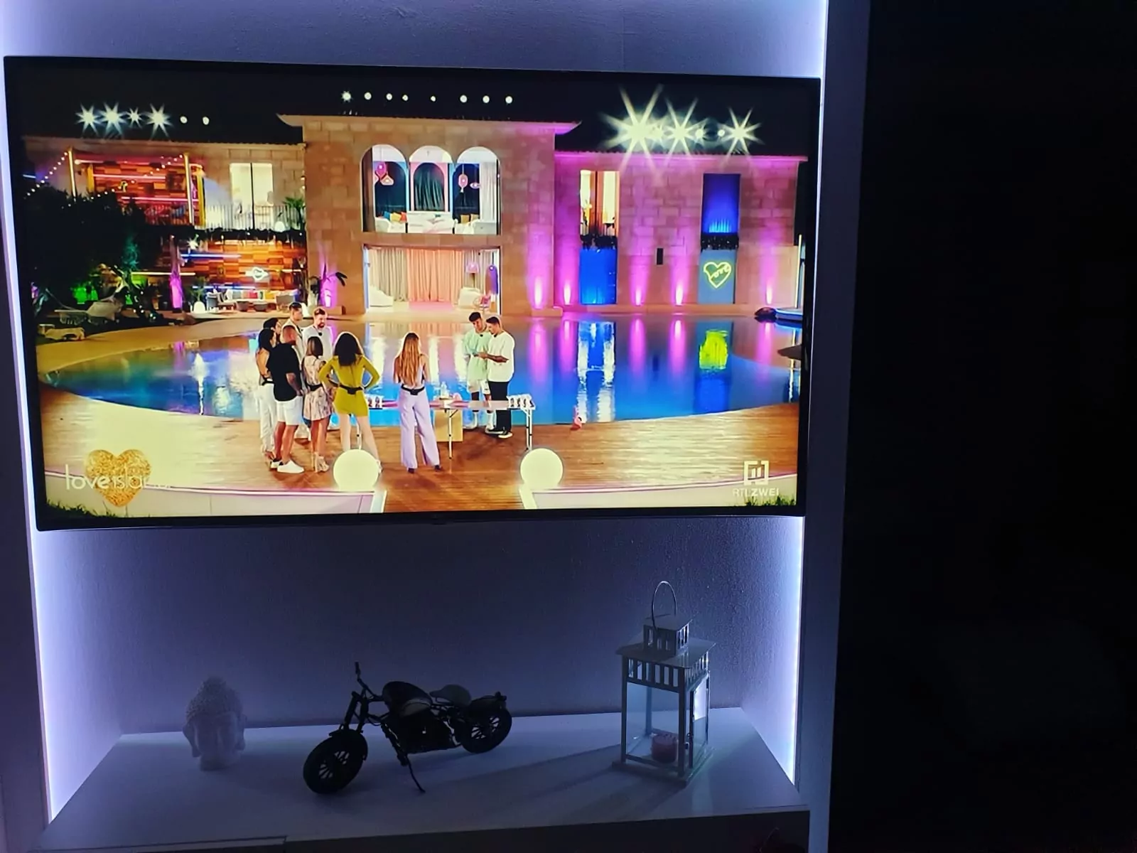 Auf einem Fernseher wird eine farbenfrohe Szene gezeigt, in der eine Gruppe von Menschen nachts an einem großen, beleuchteten Pool steht. Der Bereich ist mit leuchtenden Lichtern dekoriert und im Hintergrund ist ein malerisches Gebäude zu sehen. In der Nähe steht ein kleines Regal mit einem Motorradmodell und einer Laterne.