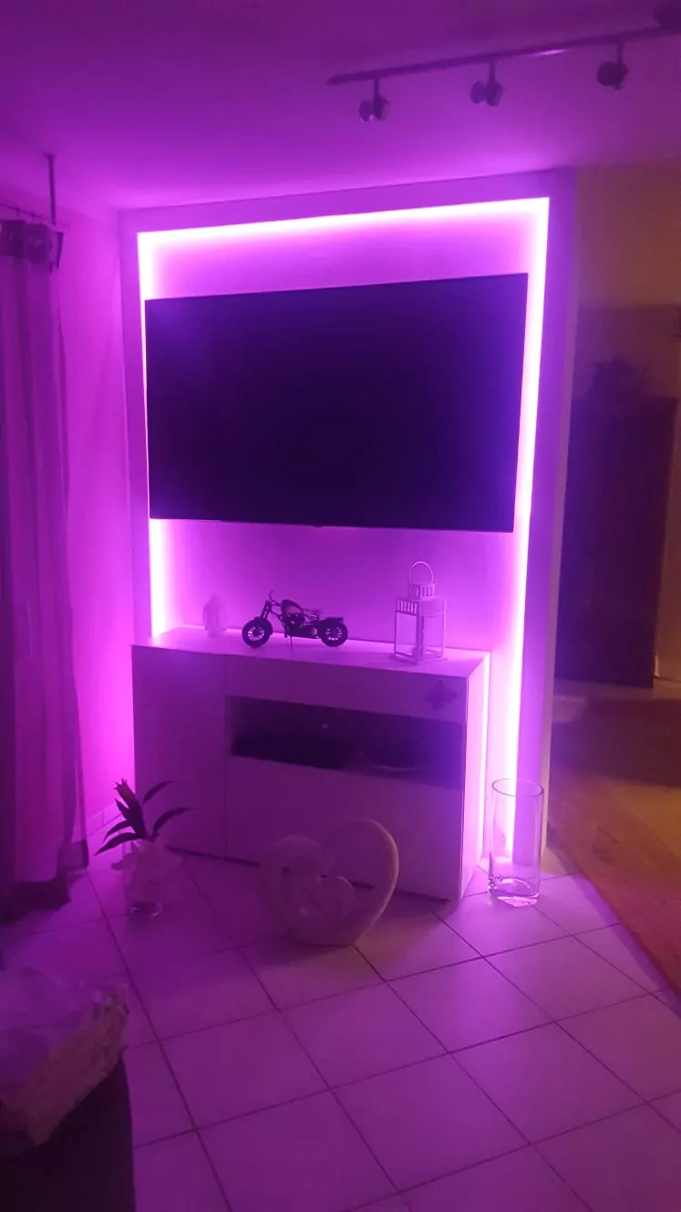 Ein modernes Wohnzimmer mit einem an der Wand montierten Flachbildfernseher, umgeben von rosa LED-Beleuchtung. Unter dem Fernseher steht ein weißer Schrank mit einem kleinen Motorradmodell und Dekoartikeln. Der Boden ist gefliest und daneben steht eine kleine Topfpflanze.