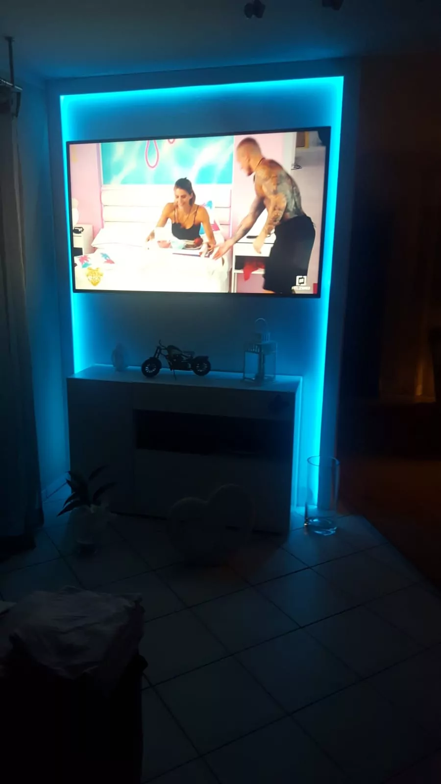 Ein an der Wand montierter Fernseher mit blauer LED-Hintergrundbeleuchtung zeigt eine Szene mit zwei Personen im Innenbereich. Unter dem Fernseher steht ein weißer Schrank mit einem kleinen Motorradmodell darauf und einigen Dekorationsgegenständen.