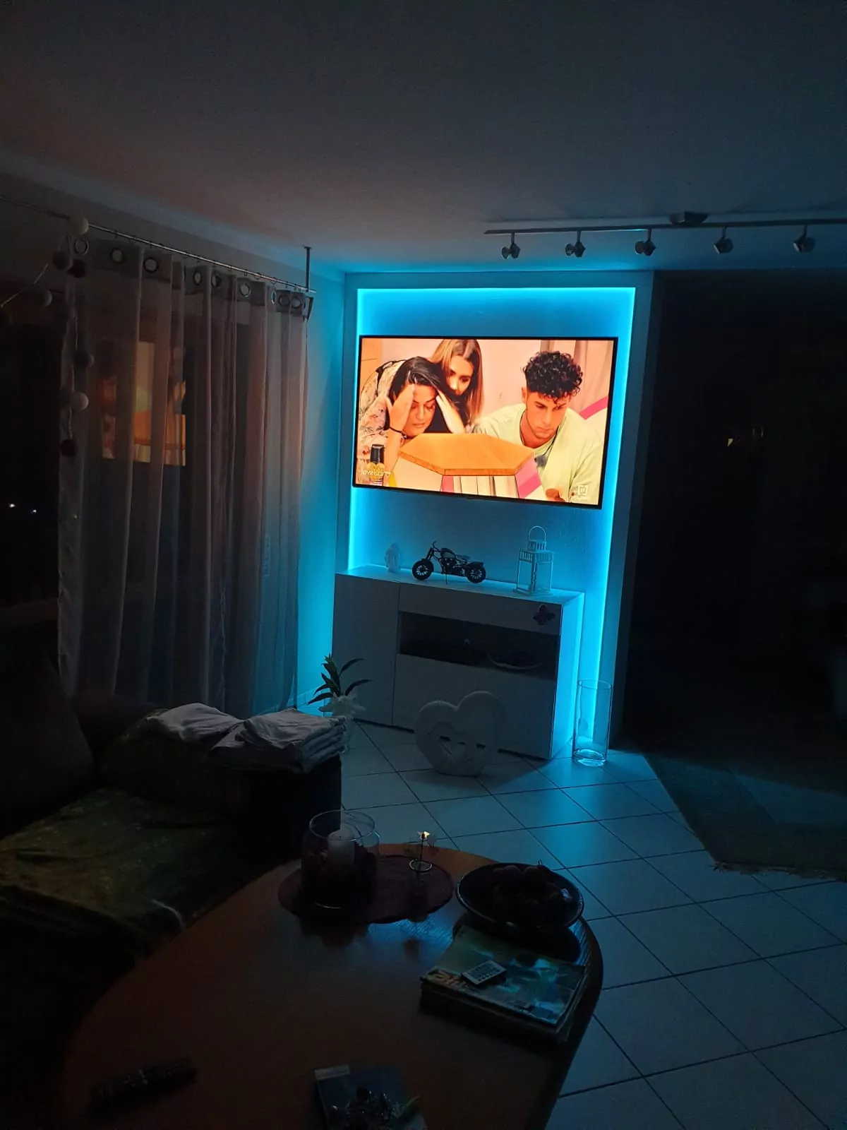 Ein Wohnzimmer, das durch die blaue Hintergrundbeleuchtung eines an der Wand montierten Fernsehers beleuchtet wird, auf dem eine Gruppe von Menschen zu sehen ist. Vorhänge verdecken ein nahes Fenster, eine kleine Pflanze und Dekorationsgegenstände stehen auf einem Regal unter dem Fernseher und im Vordergrund steht ein Couchtisch mit Büchern und Kerzen.
