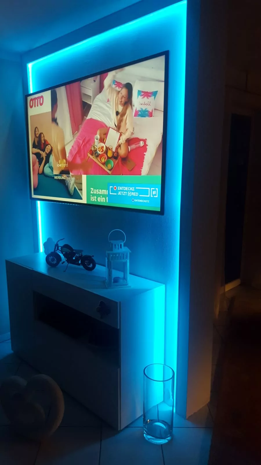 Auf einem an der Wand montierten Fernseher ist das Bild einer Person zu sehen, die auf einem Bett sitzt und ein Tablett mit Essen hält. Im Zimmer steht ein Regal mit einem kleinen Motorradmodell, es gibt eine blaue Umgebungsbeleuchtung und auf dem Boden steht eine Glasvase.