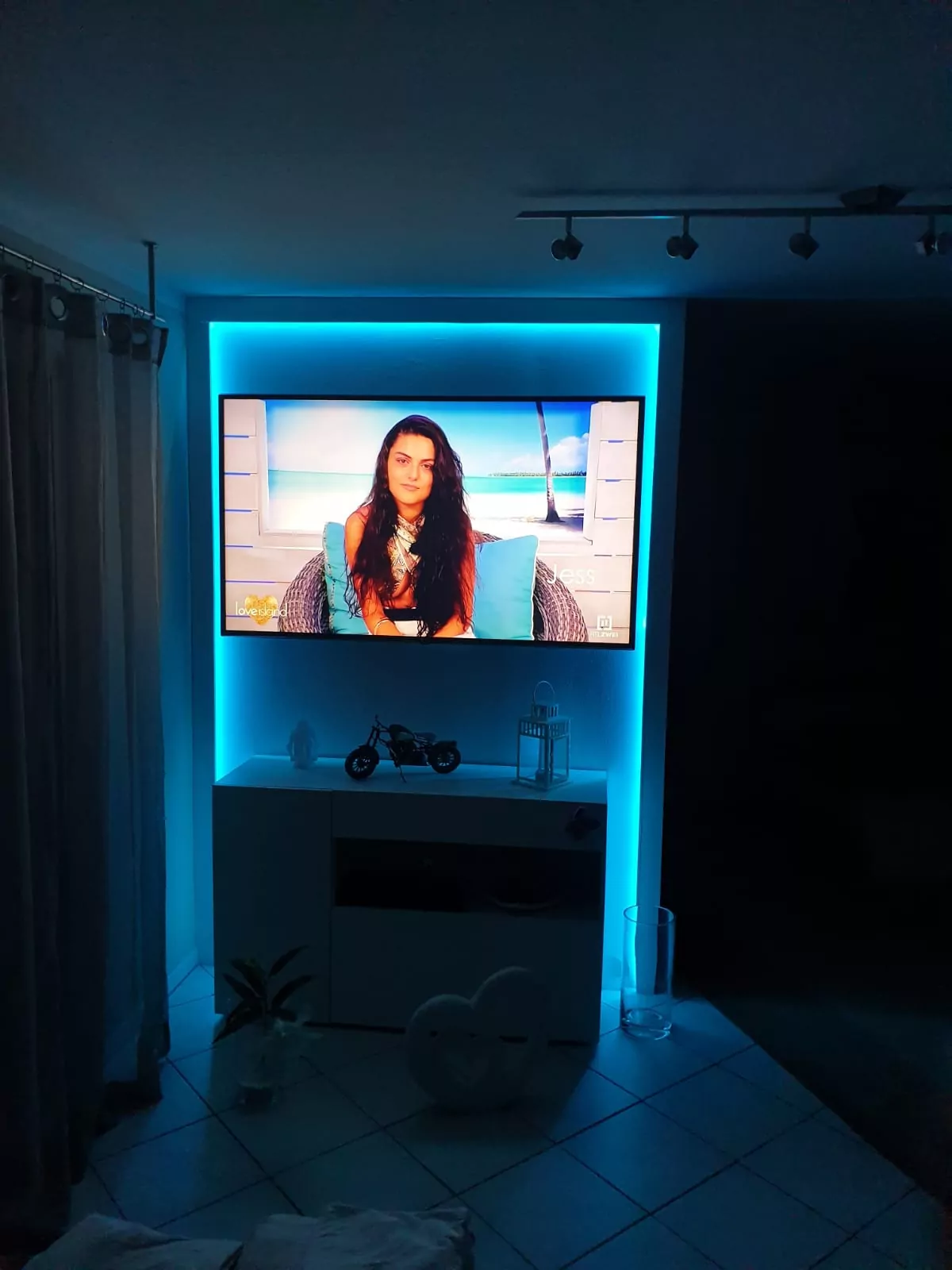 An einer Wand ist ein Fernseher angebracht, der eine Show zeigt, in der eine Person mit langen dunklen Haaren in einer Strandkulisse sitzt. Der Bildschirm ist von einem blauen LED-Licht umgeben. Unter dem Fernseher steht ein weißer Schrank mit Dekorationsgegenständen.