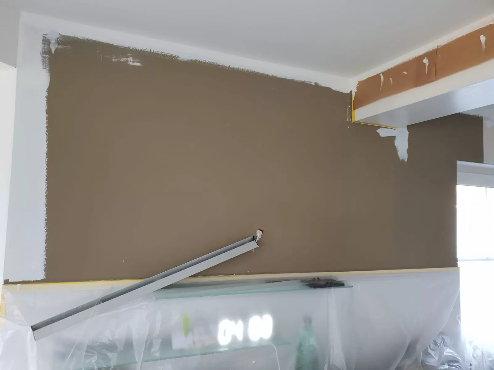 Zu sehen ist eine teilweise mit brauner Farbe und weißer Grundierung gestrichene Wand. Eine Plastikfolie deckt den unteren Bereich ab. An der Wand lehnt ein Metalllineal. Helles Sonnenlicht fällt durch ein nahes Fenster. Eine Digitaluhr zeigt „04:08“ an.