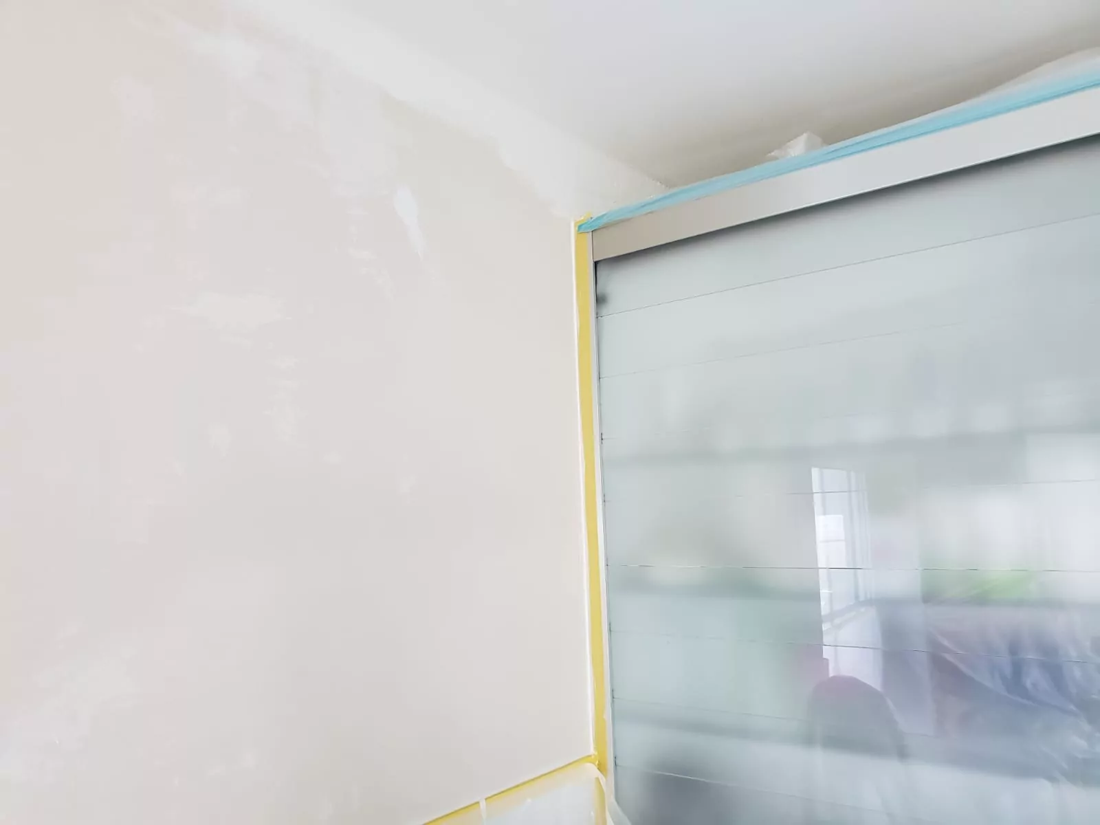 Eine teilweise bemalte cremefarbene Wand neben einer metallischen Oberfläche auf der rechten Seite. Blaues Klebeband säumt die Oberkante und gelbes Klebeband verläuft vertikal in der Ecke. Die Szene scheint ein Raum zu sein, der gerade renoviert oder gebaut wird.