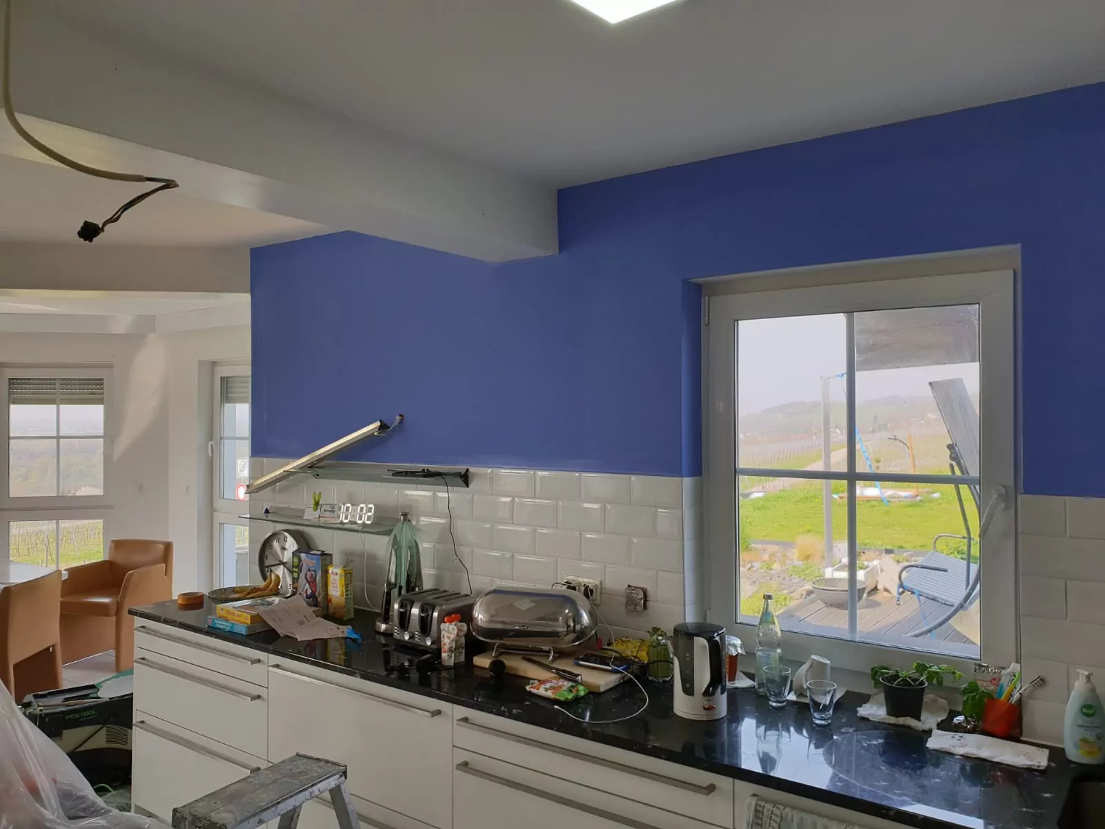 Eine Küche mit einer Arbeitsplatte, die mit Küchengeräten, Gläsern und Besteck vollgestopft ist. Die Wand über der Arbeitsplatte ist teilweise blau gestrichen. Ein Fenster öffnet sich zu einer grünen Landschaft. Unfertige Kabel und eine Trittleiter sind sichtbar.