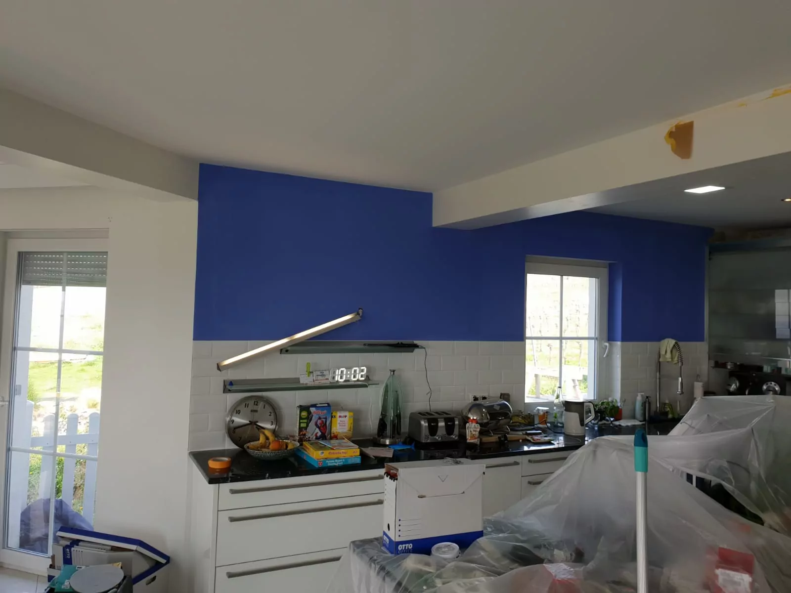 Eine Küche mit weißem Schrank und schwarzer Arbeitsplatte. Die Wand darüber ist blau gestrichen, Malerutensilien sind sichtbar. Ein Fenster lässt Tageslicht herein. Verschiedene Küchenutensilien und abgedeckte Möbel stehen herum, was auf Renovierungsarbeiten hindeutet.