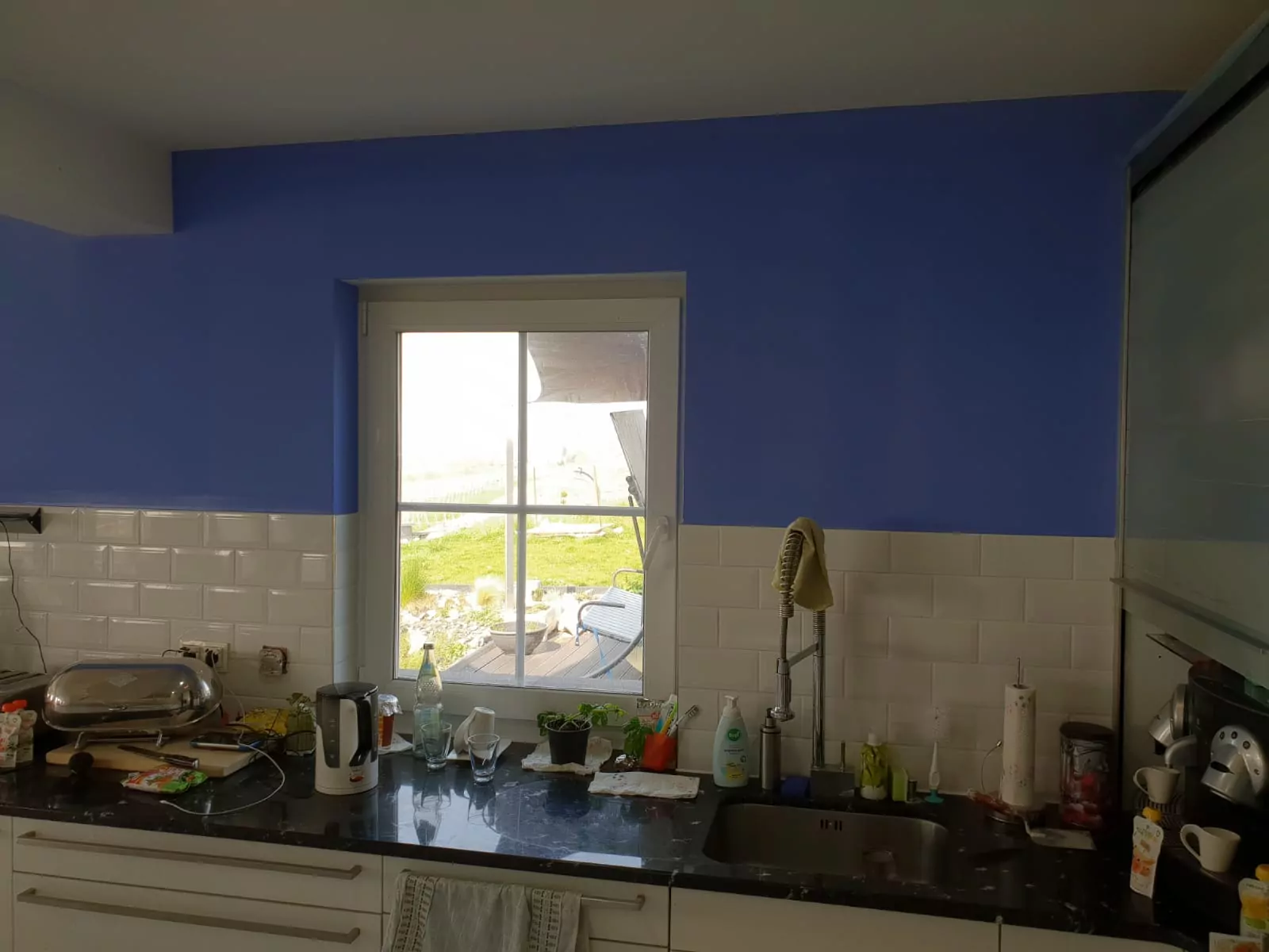 Eine Küche mit schwarzer Arbeitsplatte, Spüle und diversen Küchenutensilien. In einer gefliesten Wand mit blau gestrichenen oberen Teilen ist ein Fenster mit Blick nach draußen eingelassen. Auf der Arbeitsplatte stehen ein Wasserkocher, Obst und andere Utensilien.