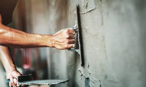 Eine Person, geschickt wie ein Maler, trägt mit einer Kelle nassen Putz auf eine Wand auf. Eine Hand hält die Kelle, während die andere eine Putzkelle hält. Die Nahaufnahme fängt die Kunstfertigkeit des Verputzprozesses ein und konzentriert sich auf geschickte Hände und präzise Werkzeuge.