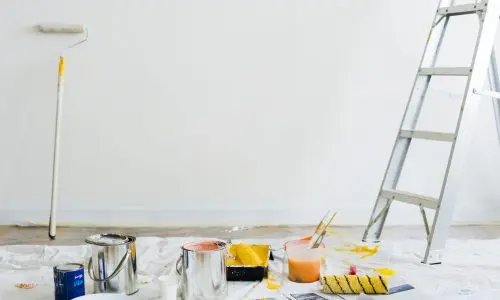 Ein Maleraufbau mit einer Leiter, Farbdosen, Pinseln und Rollen auf einer weißen Plane. Eine teilweise mit frischer weißer Farbe bedeckte Wand ist sichtbar. Eine Farbrolle lehnt an der Wand und überall liegen verstreut Zeitungen.