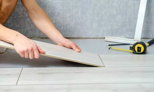 Person verlegt hellgrauen Laminatboden und richtet eine Diele an anderen bereits verlegten Dielen aus. In der Nähe liegen ein Maßband und ein Winkellineal. Im Hintergrund ist eine graue Wand zu sehen, was auf ein laufendes Heimwerker- oder Renovierungsprojekt hinweist.