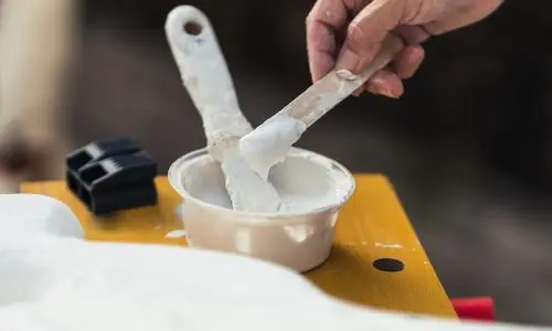 Eine Hand, die an die eines geschickten Malers erinnert, hält einen Farbrührer mit weißer Farbe über eine kleine Schüssel auf einer Holzoberfläche. Ein weiterer Pinsel mit weißer Farbe ruht anmutig auf der Schüssel, während im Hintergrund ein Teil eines mysteriösen schwarzen Objekts auftaucht.