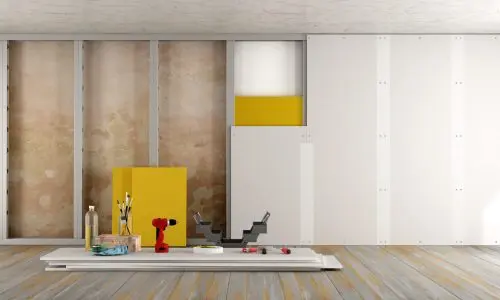 Ein Raum wird renoviert, mit Werkzeugen und Materialien. Es gibt eine unfertige Wand, einen Holzboden und Malutensilien. An der Wand steht eine gelb-weiße Tafel. Auf einem Tisch in der Nähe liegen eine rote Bohrmaschine, ein Lineal und Pinsel.