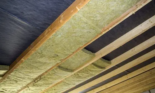 Nahaufnahme einer im Bau befindlichen Decke mit freiliegenden Holzbalken und dazwischen angebrachtem gelbem Isoliermaterial. Die Balken verlaufen parallel und die Isolierung scheint gleichmäßig angebracht zu sein, wobei einige Lücken sichtbar sind. Der Deckenhintergrund ist mit einem dunklen Stoff oder einer Barriere ausgekleidet.