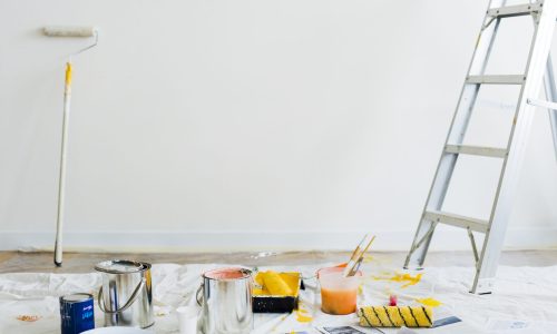 Ein Maleraufbau mit einer Leiter, Farbdosen, Pinseln und Rollen auf einer weißen Plane. Eine teilweise mit frischer weißer Farbe bedeckte Wand ist sichtbar. Eine Farbrolle lehnt an der Wand und überall liegen verstreut Zeitungen.