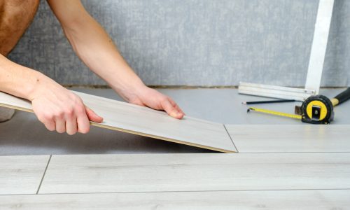 Person verlegt hellgrauen Laminatboden und richtet eine Diele an anderen bereits verlegten Dielen aus. In der Nähe liegen ein Maßband und ein Winkellineal. Im Hintergrund ist eine graue Wand zu sehen, was auf ein laufendes Heimwerker- oder Renovierungsprojekt hinweist.